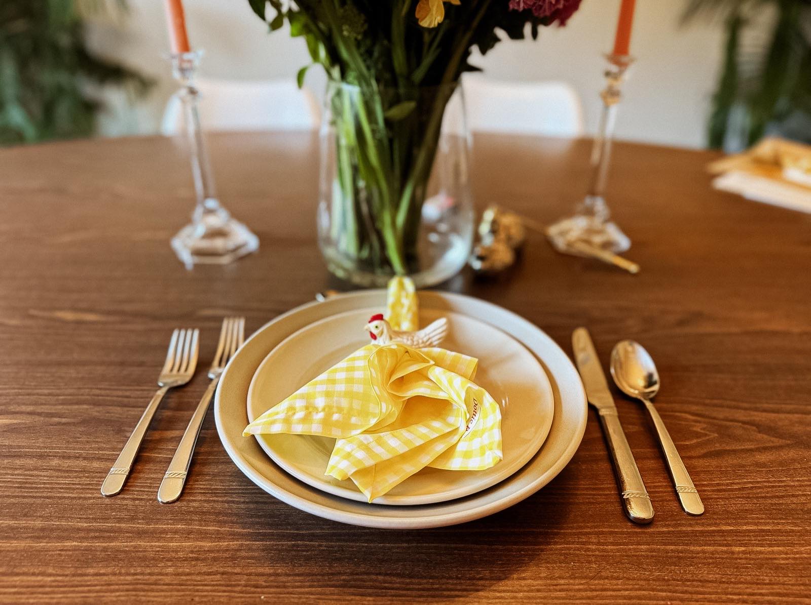 A place setting on a walnut table: a yellow gingham napkin standing in a white bowl, gold flatware either side, flowers and coral taper candles behind