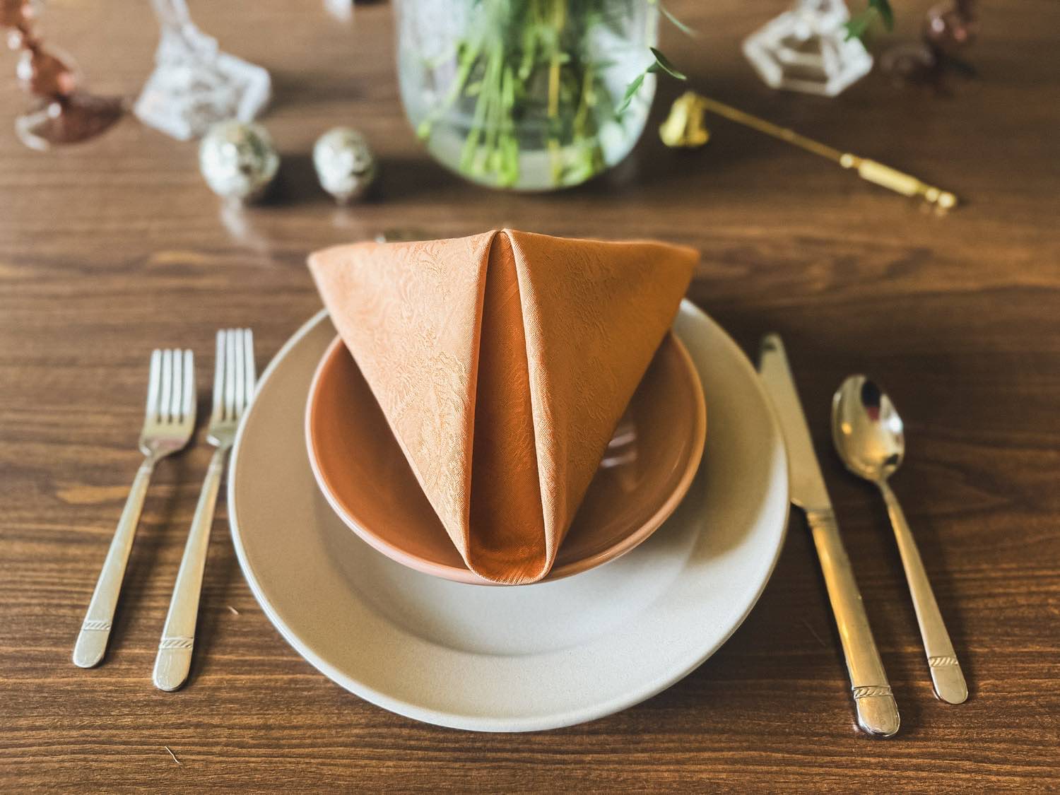 A set table with a terracotta linen napkin, gold flatware and greenery