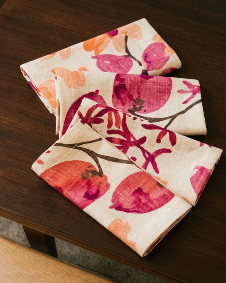Folded cream linen napkins printed with watercolour pomegranates in coral, magenta and rust, arranged on a dark walnut table